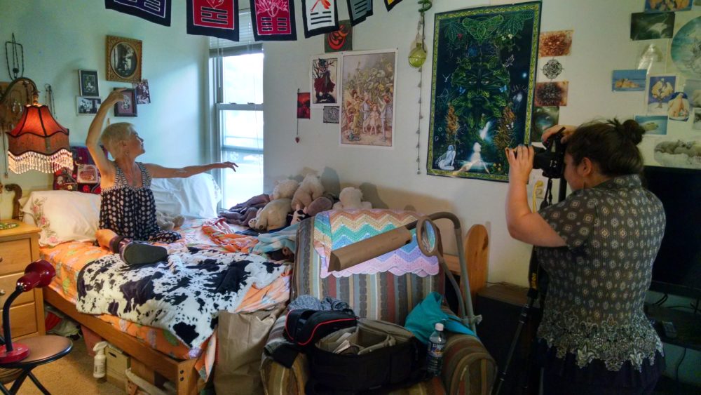 The image depicts an indoor scene where a female photographer is capturing a moment involving Lisa. Lisa is seated on a bed, pointing towards the window with a joyful expression, as if she's showing something outside. The bed is adorned with a black and white cow-print blanket and a multi-colored crochet blanket at its foot. The room is filled with personal items and decorations, including plush toys, framed pictures, and various wall hangings. The photographer, focused on her task, is standing to the right, operating a camera mounted on a tripod. There's a sense of liveliness and character in the room, reflecting the occupant's interests and personality.