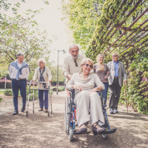 A group of six, smiling older adults walk in downtown Denver. Some people are using mobility assistance devices like a wheelchair, a cane, and a walker.