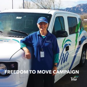 A smiling Via Paratransit driver stands in front of a MV1 accessible vehicle. The driver is an older woman who is wearing a blue Via polo, blue Via cap, and a Via badge around her neck. She is leaning on the MV1, which is white with Via's logo on the side.