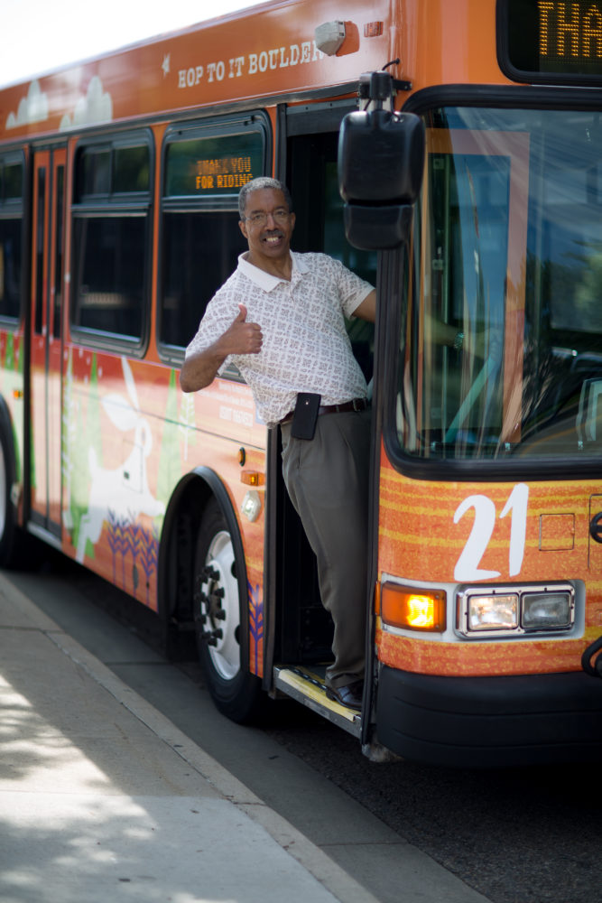 An older black man leans playfully out the open door of a HOP bus. He is smiling toward the camera and giving a thumb's up.