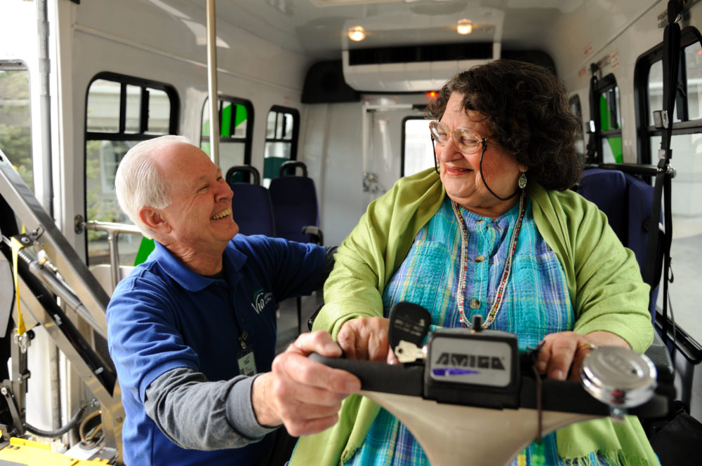 An older, white man wears a Via Mobility Services blue polo uniform shirt. He is smiling at a client, an older woman using a mobility scooter, as he secures her safely into her seat on the accessible vehicle.