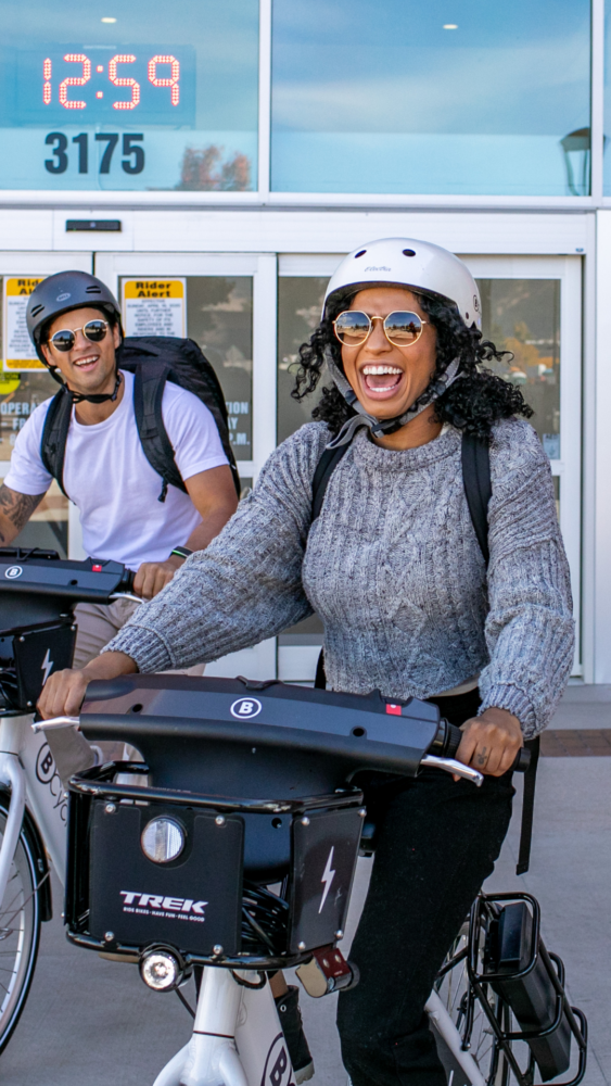 A young woman wearing a grey sweater smiles toward the camera while riding a BCycle bike. Her friend is trailing on a bike behind her. They are both wearing white BCycle helmets.