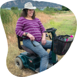 Bonnie, a Via Rider, uses her black and teal electric mobility scooter on a hiking trail in Boulder. She's wearing blue jeans, a dark and light purple striped shirt, and a white wide-brimmed hat. She's smiling at the camera.