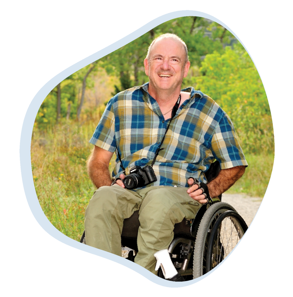 A smiling man in a wheelchair outdoors, wearing a blue and yellow plaid shirt and green pants. He has a camera around his neck and is positioned against a backdrop of greenery and trees. The setting is the South Mesa trail.