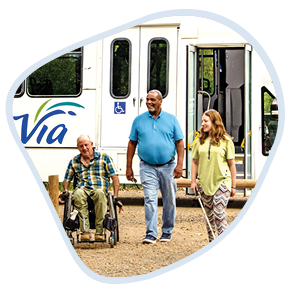 Three people outdoors: an older man using a wheelchair, Topher, a man in a blue polo shirt standing next to him, Kevin, and a young woman in a yellow top and patterned pants, Sarah, walking beside them. They are near a white bus with the logo "Via" on it. The setting is the parking lot of a park.