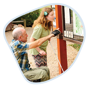 The same young woman, Sarah, looks closely at a trail map at the South Mesa Trailhead in Boulder. Her friend, an older man in a blue and yellow checked shirt and sitting a wheelchair, points to the map. The image is shaped in a rounded frame with a light blue border.