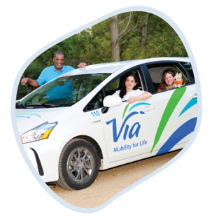 Kevin wears a blue polo shirt. He's standing next to a white car labeled "Via - Mobility for Life". Inside the car, a woman in the driver's seat and a young woman in the passenger seat are both looking out of the open windows, smiling. The setting appears is an outdoor area with trees in the background.