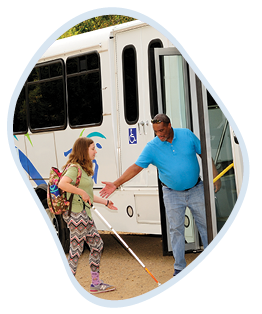 Kevin in a blue polo shirt holding open the door of a white bus, gesturing to assist Sarah, wearing a patterned outfit and a backpack. Sarah, who is holding a white cane, is stepping towards the bus. The environment suggests an outdoor setting.
