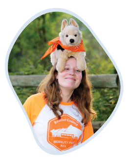 Sarah wearing an orange t-shirt with the text "Boulder County - Mobility for All" smiles with her eyes partly closed. Atop her head is a plush toy dog wearing an orange bandana. The image is presented within a rounded frame with a light blue border.