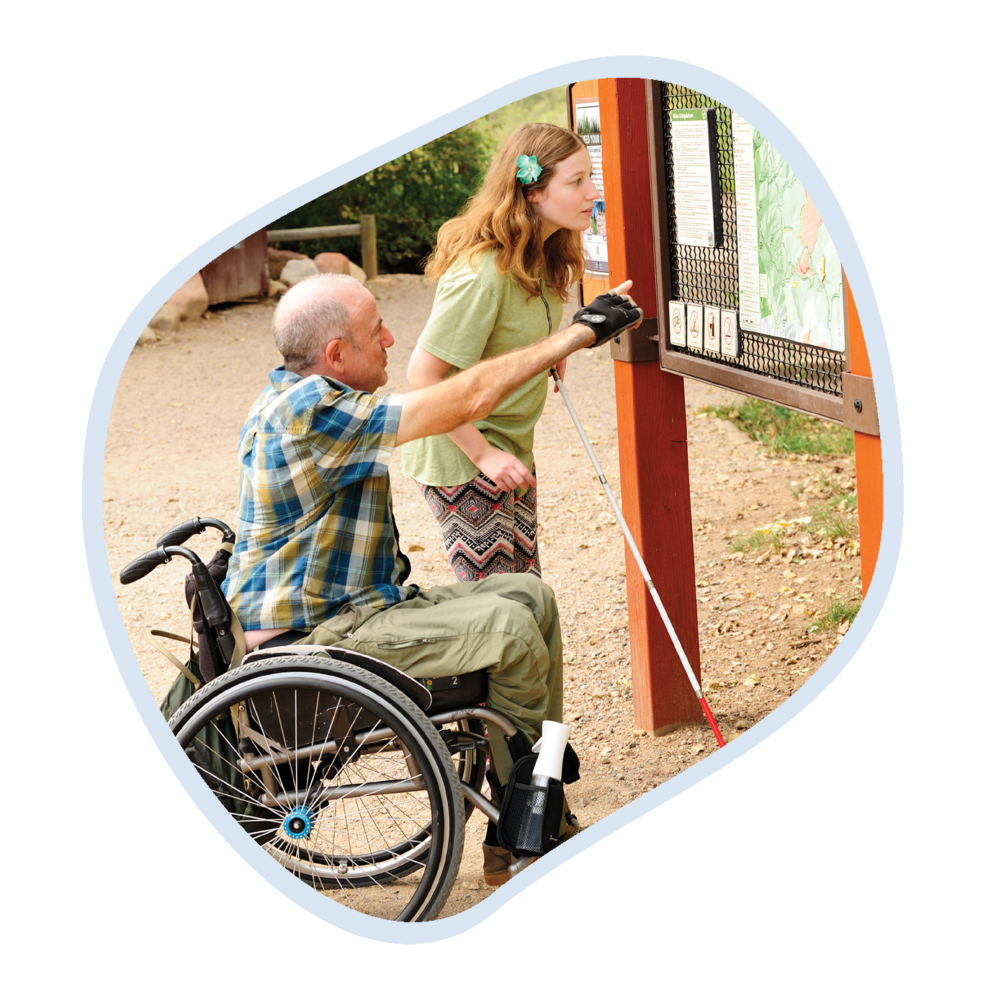 A man in a wheelchair and a young woman, both outdoors, interacting with an information board. The man, wearing a blue plaid shirt and green pants, points to the board. The woman, in a green shirt and patterned pants, leans in to get a closer look. They are at the South Mesa trailhead.