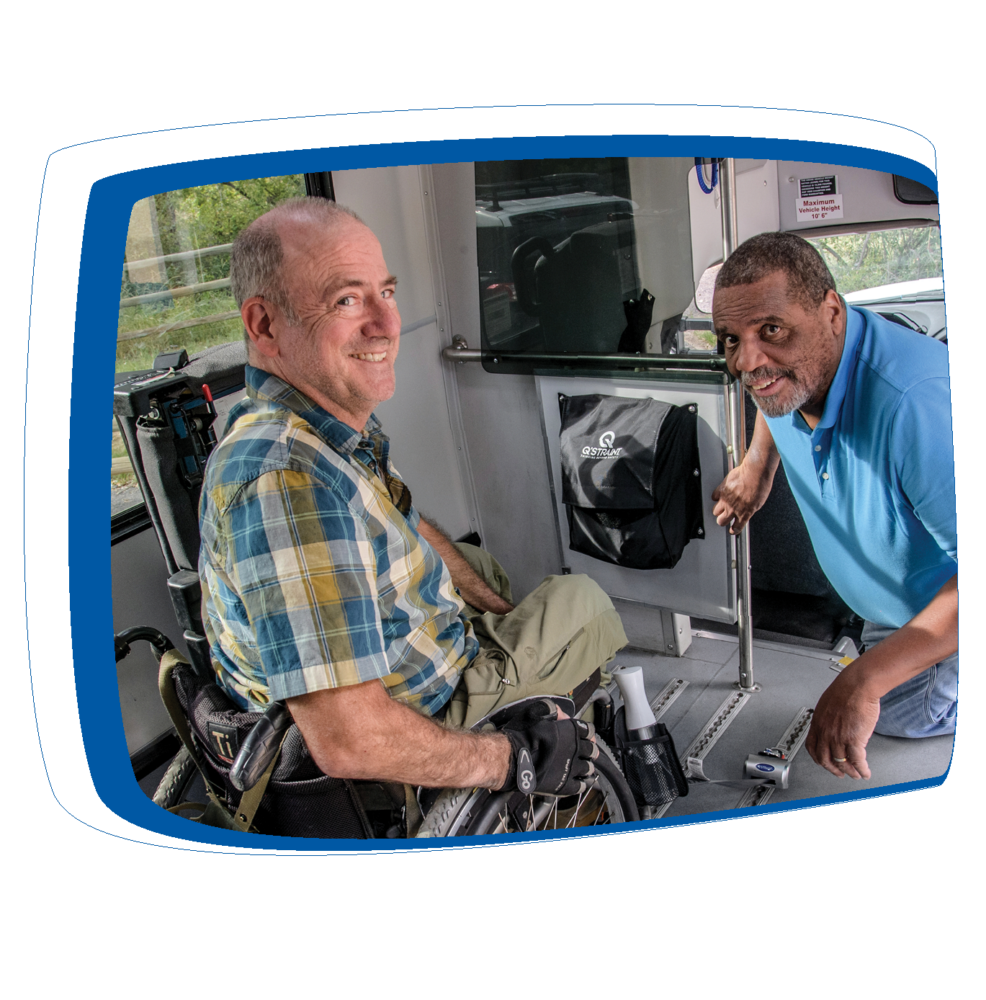 Two men inside a vehicle, one seated in a wheelchair wearing a blue and yellow plaid shirt with a camera on his lap, and the other man, wearing a blue polo shirt, leaning over assisting with the wheelchair's positioning. The interior of the vehicle has designated areas for wheelchair accommodation and a bag with a logo hangs on the side wall. Both men are smiling, and a window provides a view of the outdoors.