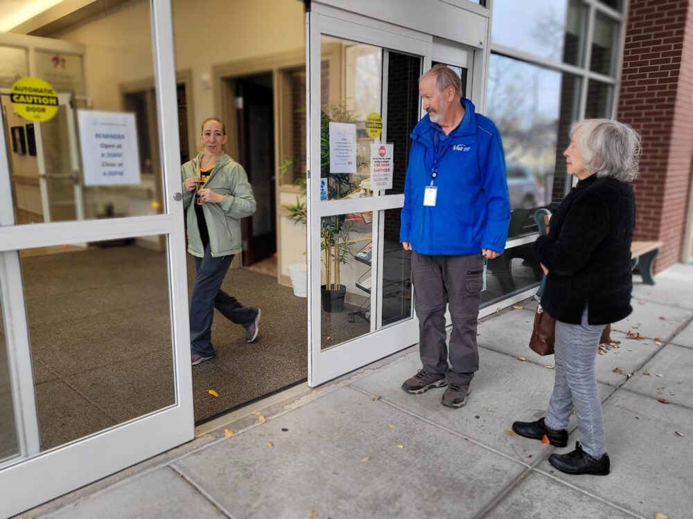 Outside Seniors Resource Center, a building with automatic glass doors, three people are engaged in a conversation. On the left, a staff woman exits the building; she has shoulder-length hair, wears a pale green jacket, holds a yellow mug, and is stepping forward. In the center stands a male Via driver in a blue jacket with a badge, facing the woman to his left. To his right is Suzy, an older woman with shoulder-length gray hair, wearing a black cardigan and holding a brown purse. The entrance has a "CAUTION AUTOMATIC DOOR" sign and posted operating hours. Fallen leaves on the sidewalk suggest it is autumn.
