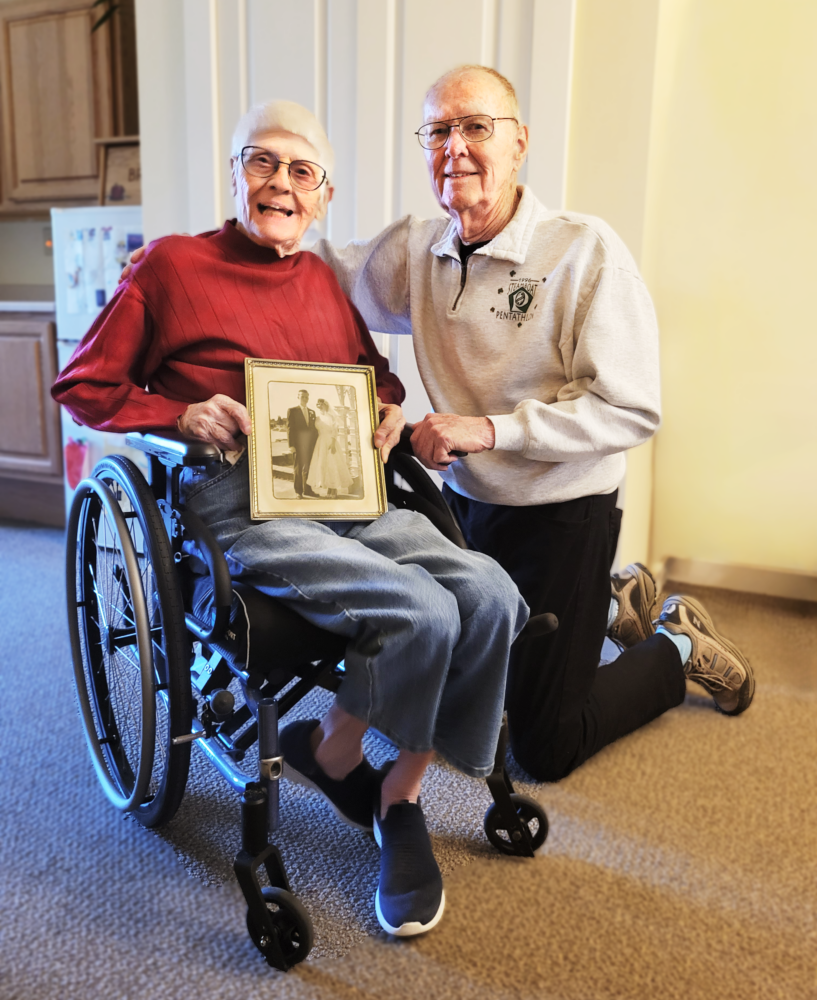 An elderly couple, Doreen and Ray, are smiling in an indoor setting, Doreen is seated in a wheelchair and Ray is kneeling beside her. Doreen is wearing a red sweater, blue jeans, and black shoes, while holding a framed black-and-white photograph of the couple. Ray is wearing a gray sweater with a logo, black pants, and gray sneakers. There's a kitchen area in the background.