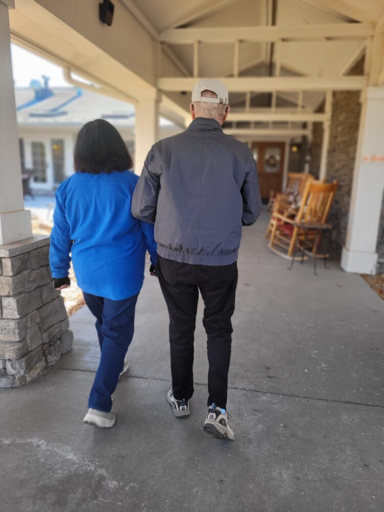 Ray is assisted by Via driver Mary as they walk side by side toward the assisted living facility where Doreen resides. It's a building with a covered entrance. Ray is wearing a gray jacket, black pants, and gray sneakers with blue accents. He has a white cap on his head. Mary is wearing a blue jacket and navy blue pants with white shoes. It's a calm and clear day.