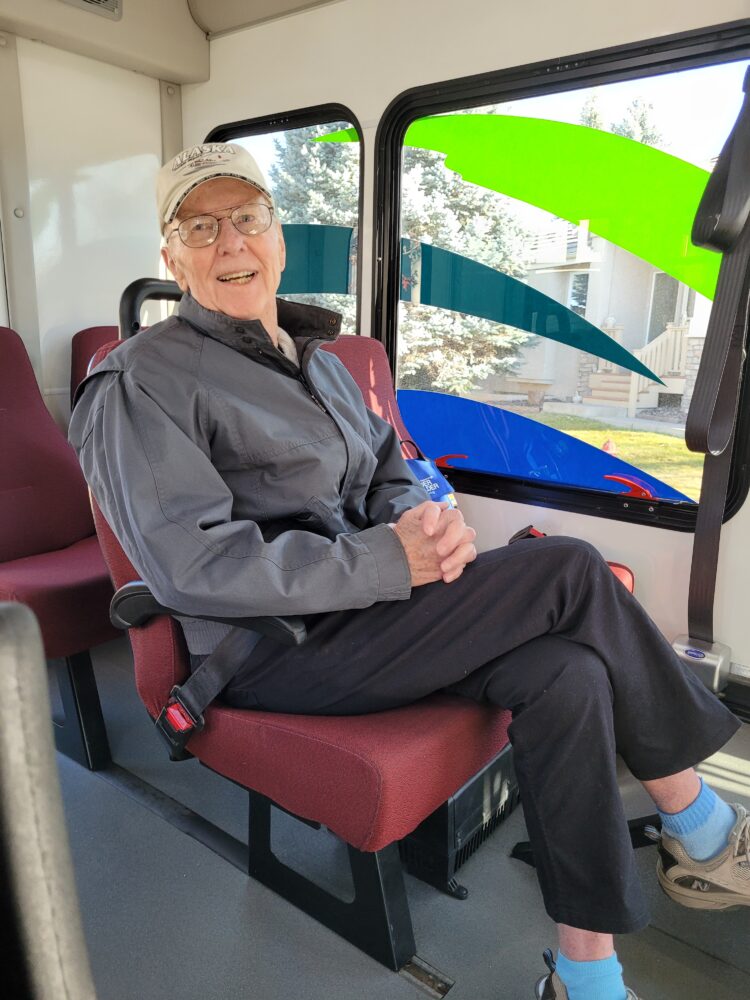 Ray is seated on a red bus seat, buckled in with a seat belt, and smiling at the camera. He is wearing a gray jacket, black pants, light blue socks, and gray sneakers. He sports a white cap with the word "ALASKA" on it. The bus interior is visible, with bright sunlight coming through the windows, and the exterior environment reflects suburban scenery.