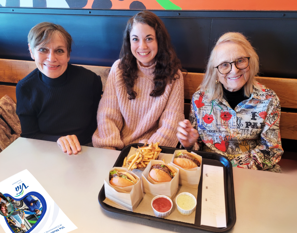 Three women of varying ages are seated at a restaurant table smiling towards the camera. On the table, there's a tray with two burgers, a pile of fries, and three containers of condiments. In the foreground, there's a flyer with the logo of Via Mobility Services, indicating a casual or promotional event. The eldest woman on the right wears a colorful jacket with artistic designs, while the other two are in more subdued attire.