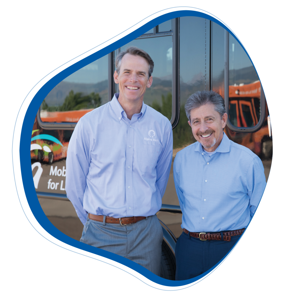 The image features Frank and Chris, two smiling men standing in front of a bus with a partial view of a banner that reads "Mobility for Life". Chris, on the left, is taller, wearing a blue button-up shirt with a logo that says "Alpine Bank", and grey pants. Frank is on the right and is wearing a blue button-up shirt with a pattern, grey pants, and a brown belt. Both appear cheerful and are looking at the camera. The bus in the background has orange, green, and white colors with a mountain graphic, which is likely part of Via Mobility Services' fleet, given the visible part of their tagline. The image is encased in a blue circular border, suggesting it might be used for marketing or informational content.
