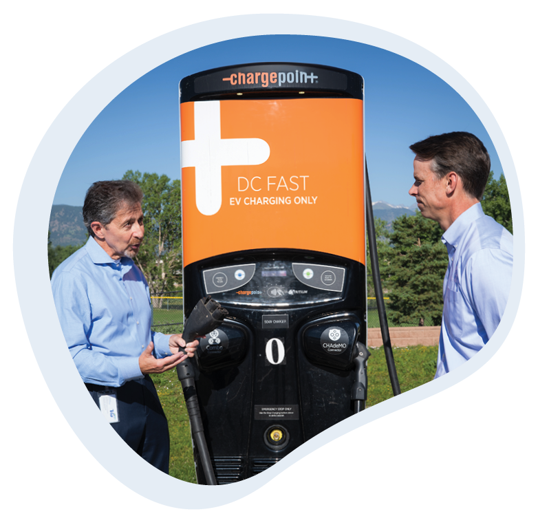 The image shows two individuals, Via CEO Frank W Bruno and Alpine Bank Market President Chris Maughan, standing in front of a ChargePoint DC fast electric vehicle charging station. Frank is on the left and holding a charging plug and appears to be demonstrating or explaining how to use the charger. Both are engaged in a conversation and dressed in business casual attire. The backdrop includes clear skies and some greenery outdoor. The charging station is prominently colored with black and orange, and the words "DC FAST EV charging only" are visible.